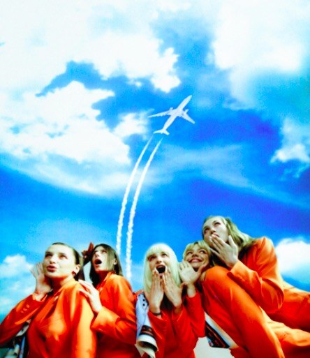 Cabin crew in orange uniforms looking up at a plane in the sky